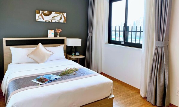 A hotel room with a bed, a nightstand, a lamp, a clock, a window with curtains, and a city view. - Roygent Parks Hanoi (Hanoi)