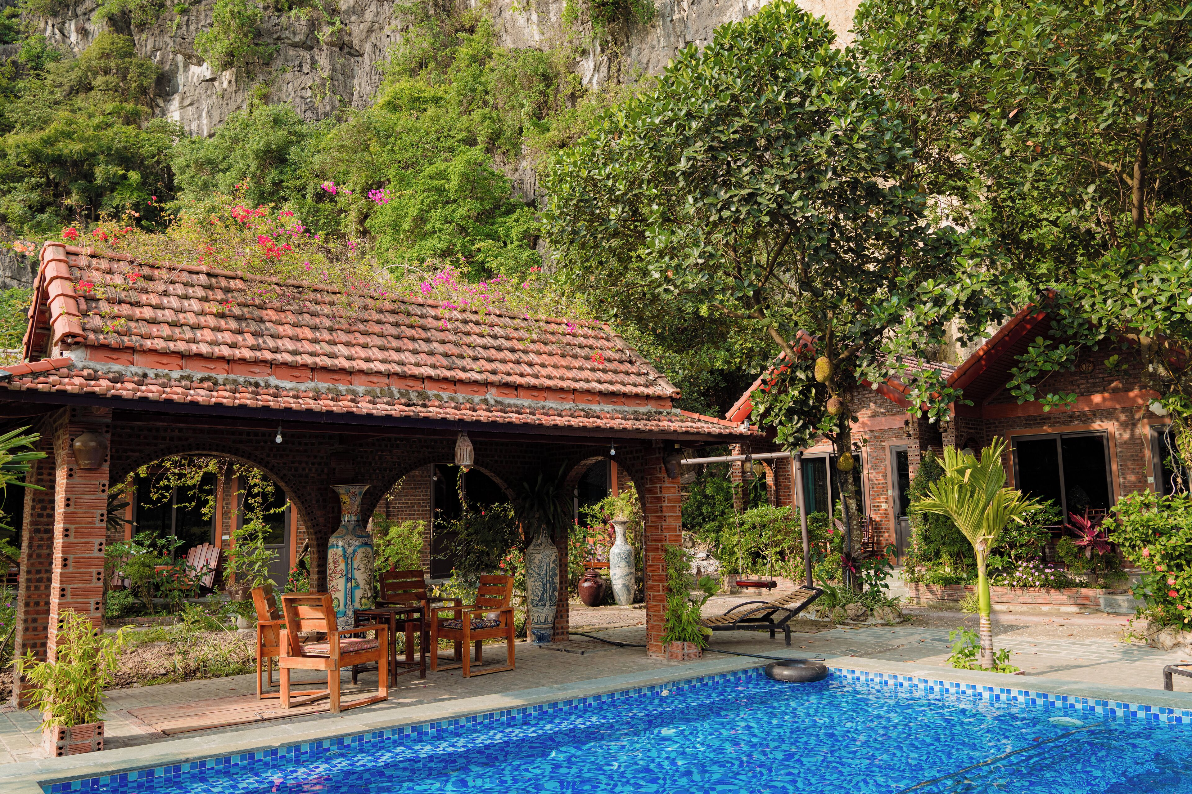 A pool area with a tiled deck, a covered seating area with chairs, and a view of a mountainous landscape.