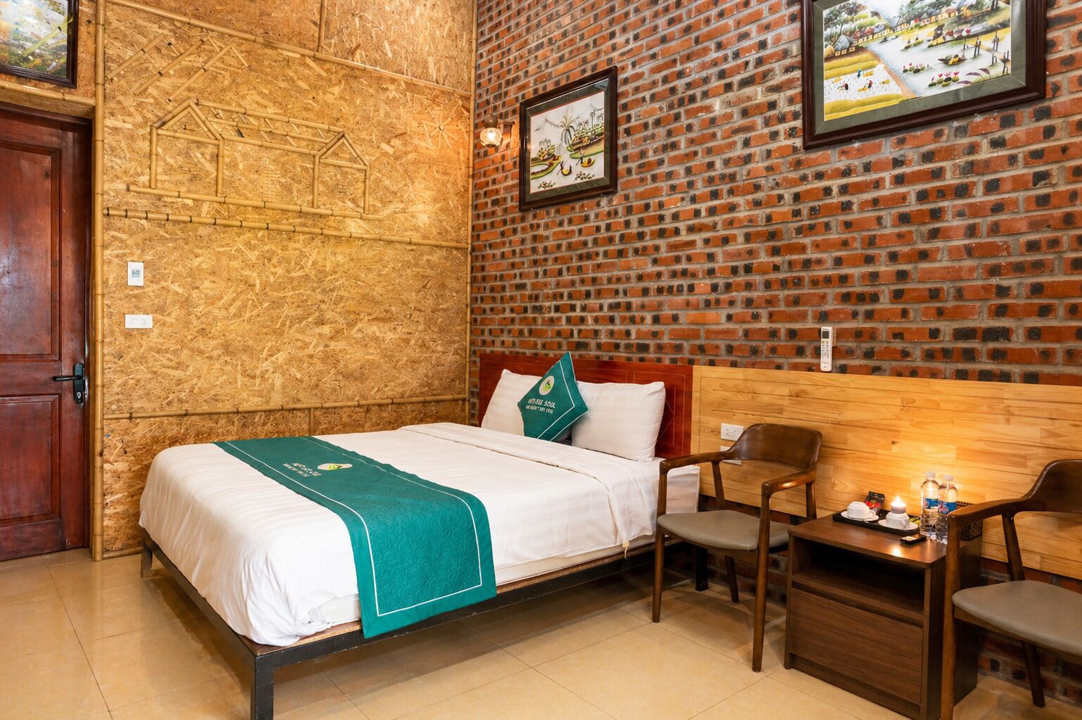 Une chambre d’hôtel avec un lit, une tête de lit en bois, un bureau avec une chaise et un mur en briques orné de tableaux.