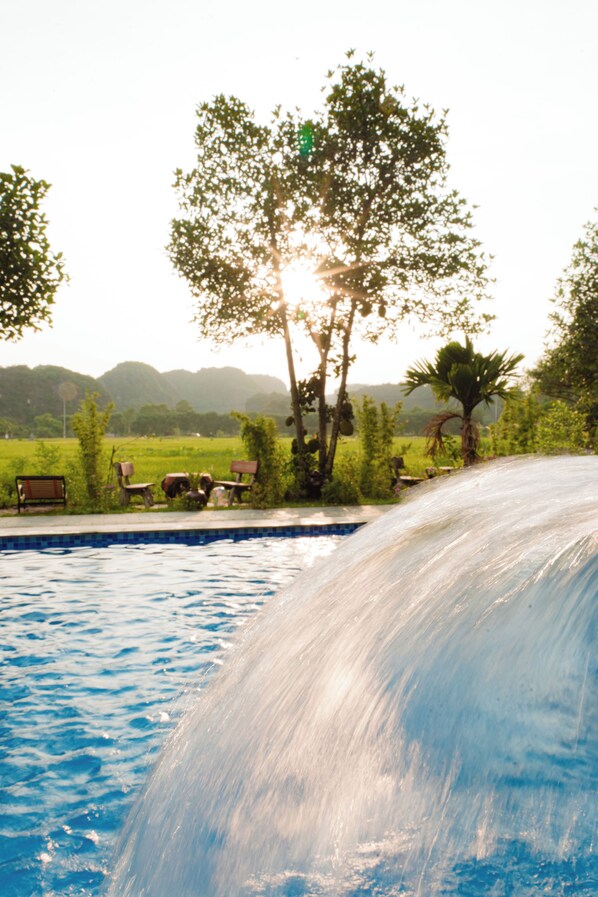 A swimming pool with a waterfall feature, surrounded by outdoor seating and palm trees. - Heritage Soul Bungalow Tam Coc (Nam Hoa Lu)