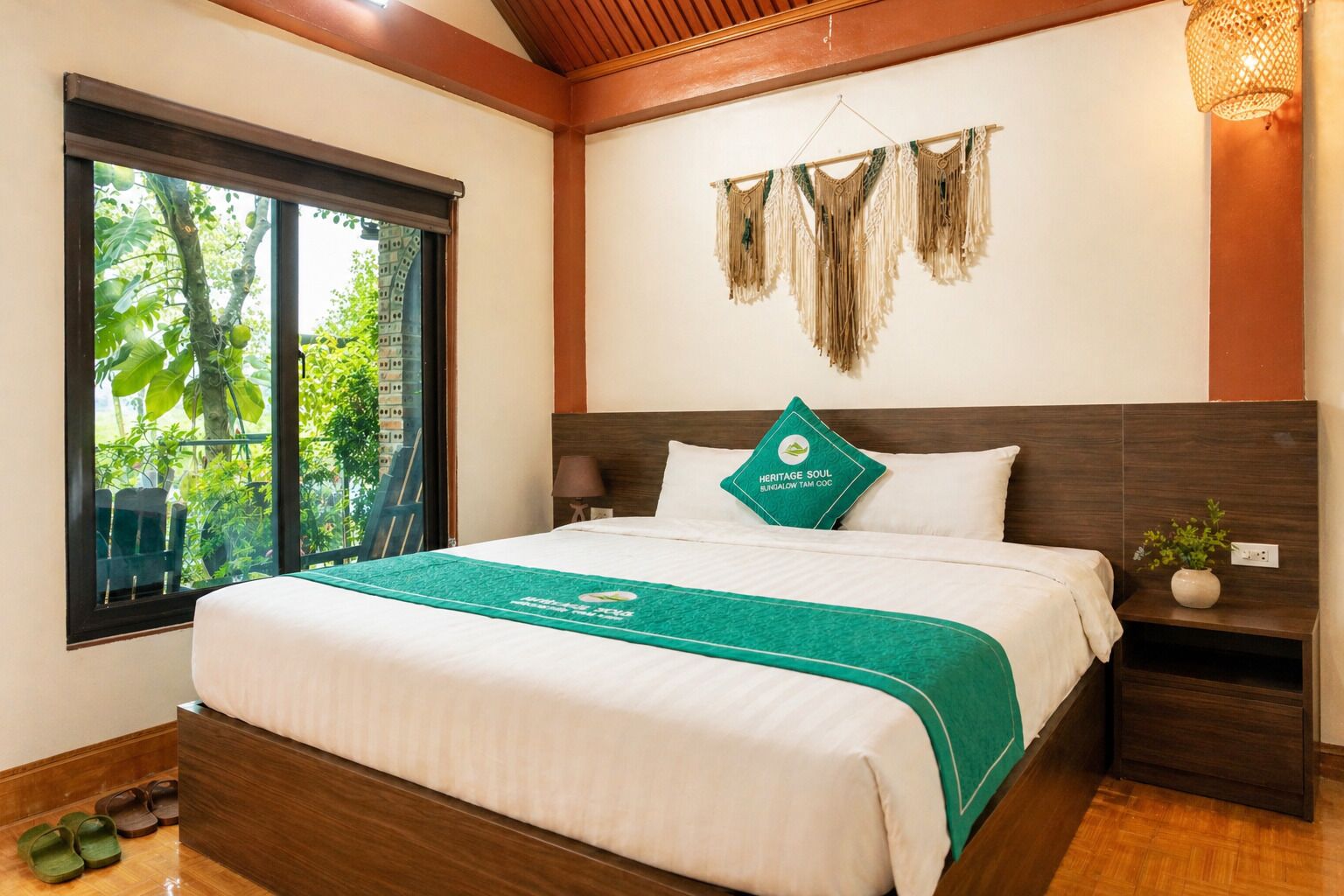 A hotel room with a large bed, a nightstand, a lamp, a window with a view of greenery, and a decorative wall hanging.