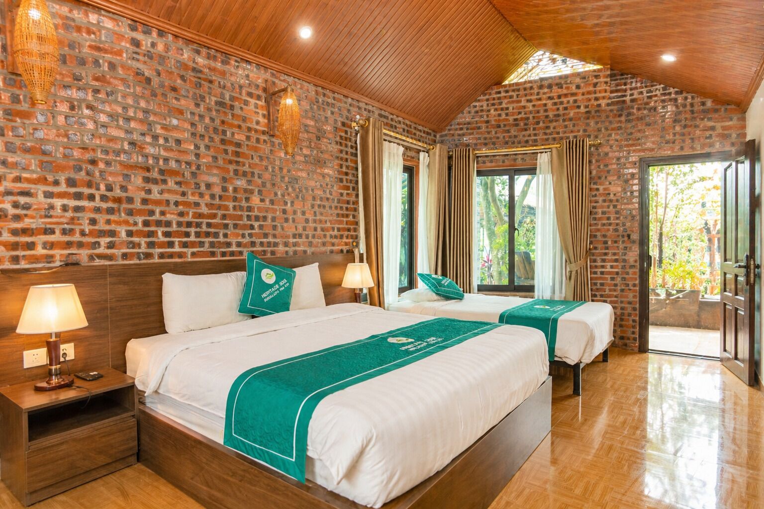 Une chambre d’hôtel avec un mur en briques, un plafond en bois, deux lits recouverts de couvre-lits verts, une table de chevet, une lampe et une fenêtre avec des rideaux.