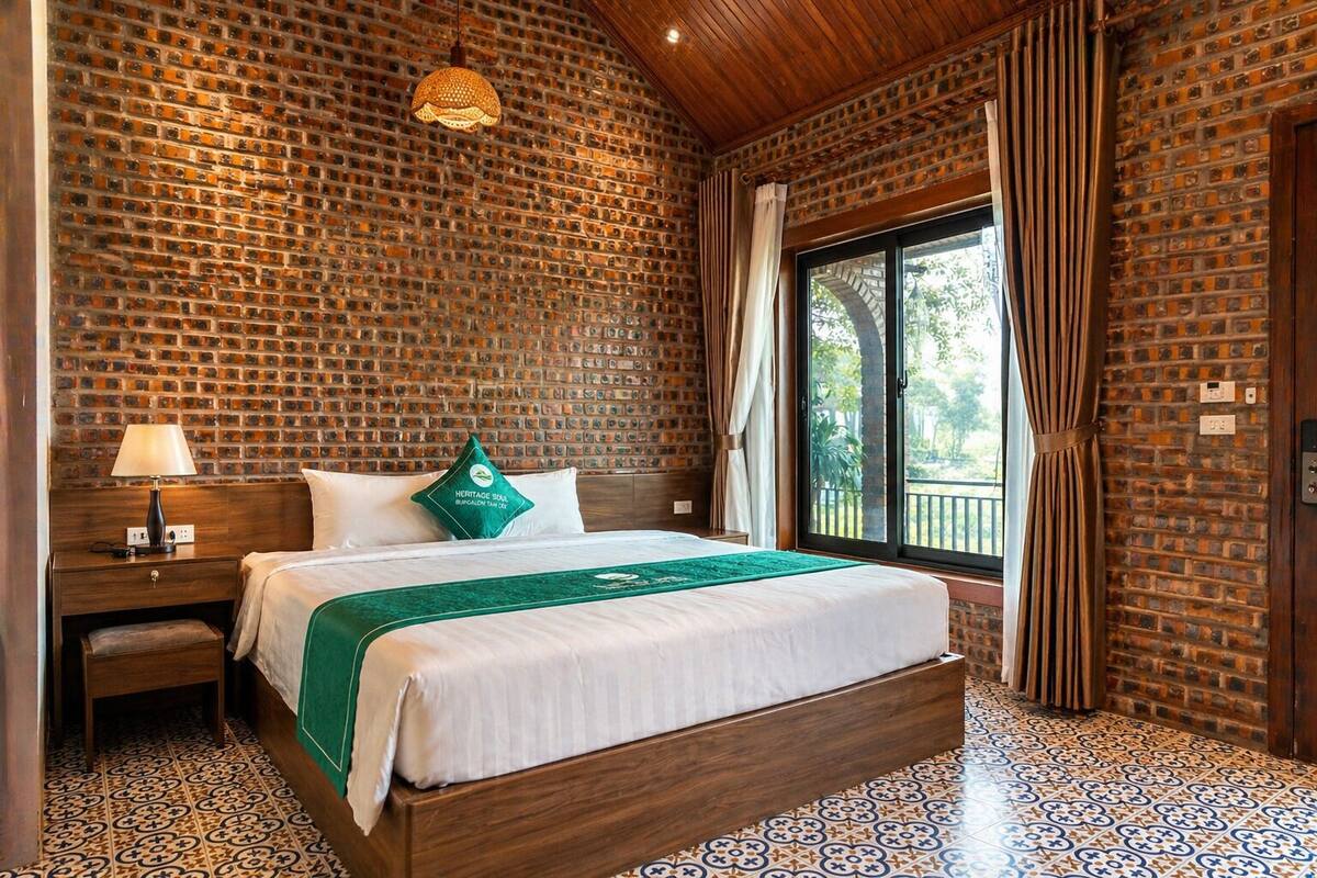 Une chambre d’hôtel avec un mur en briques, un lit recouvert d’un plaid vert, une table de chevet, une lampe et une fenêtre avec des rideaux.