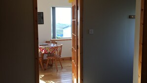 Um quarto com piso carpeteado, uma porta de madeira que dá para uma sala de jantar com mesa e cadeiras, e uma janela com vista para o exterior.