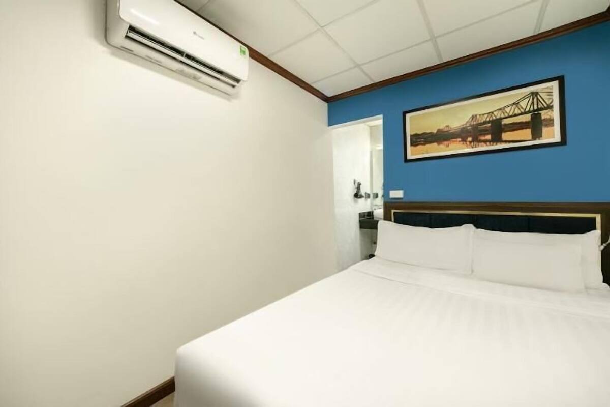 Habitación de hotel con cama, unidad de aire acondicionado, pintura de un puente y puerta que da a un baño.