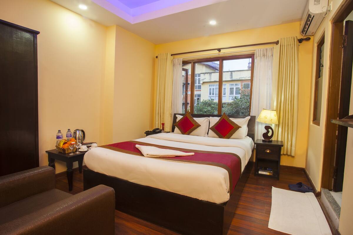 A hotel room with a large bed, a bedside table with a lamp, a chair, and a window with curtains.