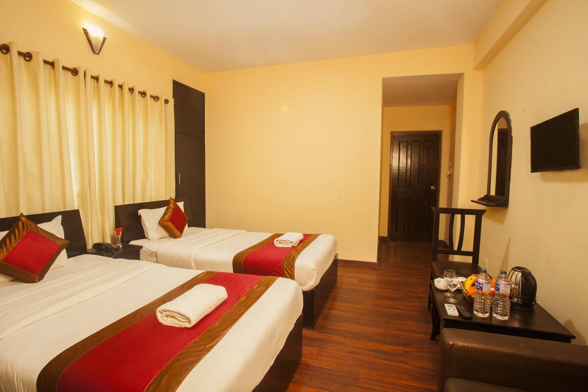 A hotel room with two beds, each with a red and white bedspread, a wooden headboard, and a small table with a remote control, water bottles, and a coffee pot.