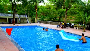 A swimming pool with people in it, surrounded by trees and a building in the background. - Hiranya Resorts (Chhatrapati Sambhajinagar)