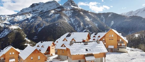 A cluster of wooden cabins with snow-covered roofs, set against a backdrop of snow-capped mountains and a clear sky.
