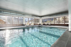 Ein heller, großer Innenpool mit klarem Wasser, Panoramafenstern und Liegestühlen.