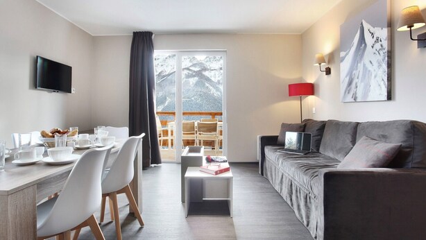A modern living room with a grey sofa, a dining area with a table set for two, and a large window with a view of snow-covered mountains. - Chalet in Praroustan with Mountain Views (PRA LOUP 1500)
