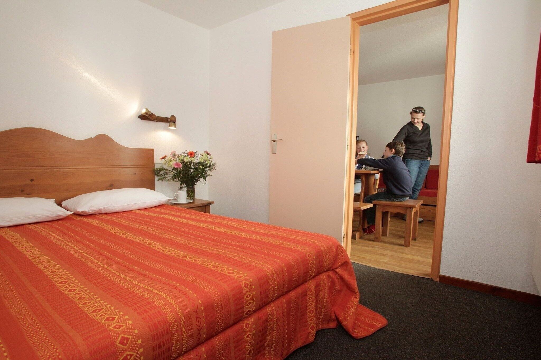 A hotel room with a bed, a nightstand with a vase of flowers, a wooden headboard, and a door leading to a dining area.