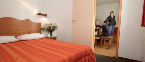 Ett hotellrum med en säng, ett nattduksbord med en vas med blommor, en träpanel som huvudgavel och en dörr som leder till en matplats.