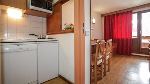 Eine kleine Küche mit Mikrowelle, Herd und Waschmaschine. Durch einen offenen Durchgang ist ein Essbereich mit Holztischen und einem Tisch zu sehen.