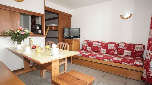Ein gemütlicher Wohnbereich mit einem Holztisch, der für das Frühstück gedeckt ist, einer Couch mit rot-weißer Musterdecke und einem Fernseher auf einem Holzboden.