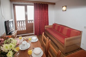 A room with a wooden table set for breakfast, a red sofa, and a television on a wooden stand.
