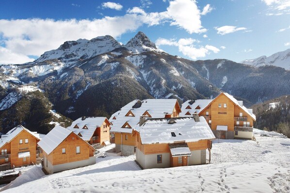 A cluster of wooden cabins with snow-covered roofs, set against a backdrop of snow-capped mountains and a clear sky.