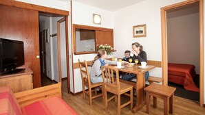 A family dining in a room with a wooden table, chairs, a television, and a red bed. - Chalet-Style Stay in Val Claret (Tignes)
