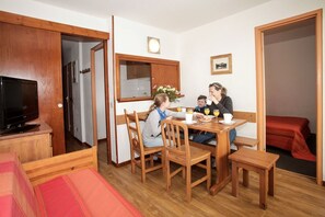 Eine Familie isst in einem Raum mit einem Holztisch, Stühlen, einem Fernseher und einem roten Bett.