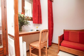 Ein Zimmer mit einem Holztisch, einem Stuhl, einem roten Sofa und einem Fenster mit roten Vorhängen.