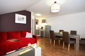 A compact living space with a red sofa, a dining table with chairs, a kitchenette, and a ceiling light fixture. - Alpine Stay with Balcony (Auris en Oisans)