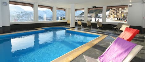 An indoor swimming pool with clear blue water, surrounded by lounge chairs and large windows offering a view of snowy mountains.
