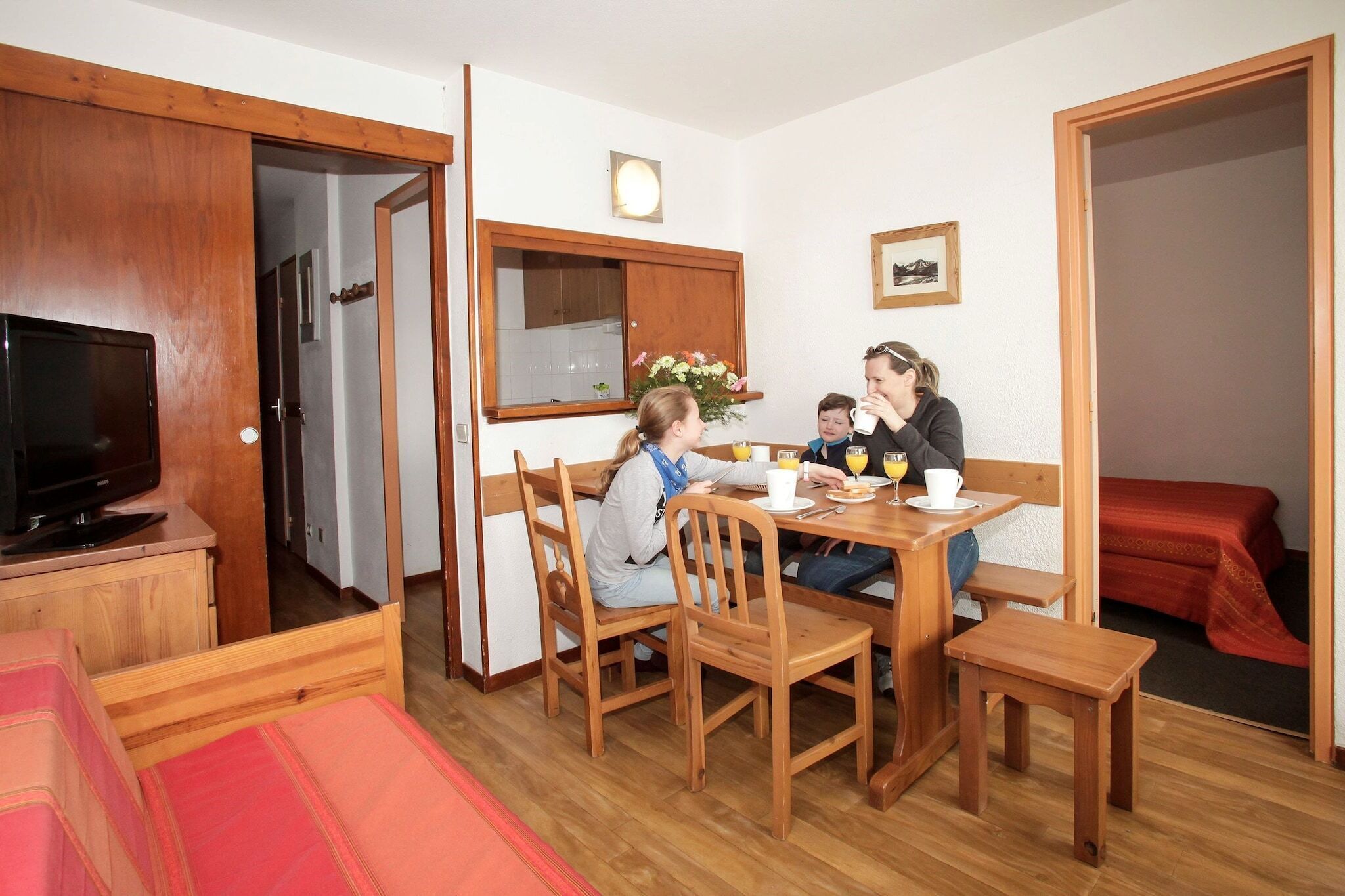 A family dining in a room with a wooden table, chairs, a television, and a red bed.