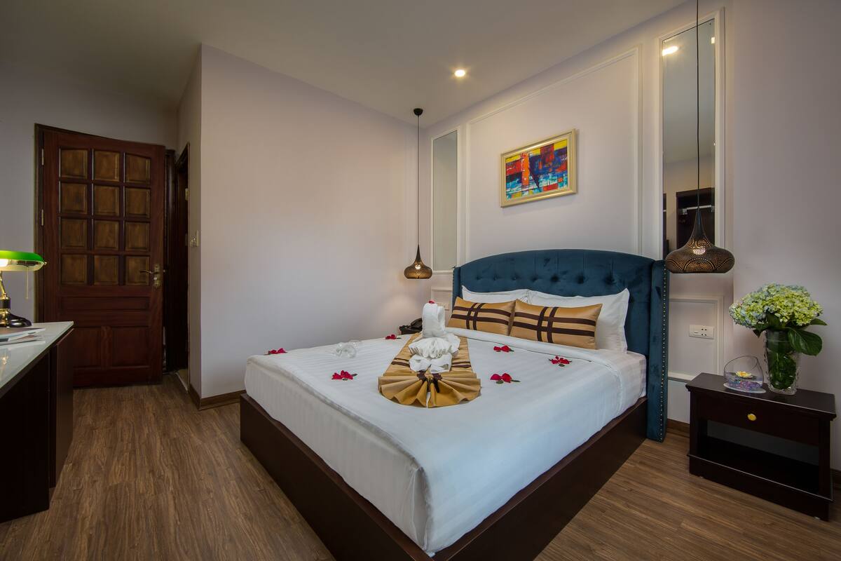 A neatly made bed with a blue headboard, a bedside table with a vase of flowers, and a painting on the wall.