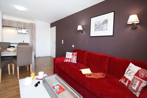 A modern living room with a red sofa, a dining area with a table and chairs, a kitchen with a stove, and a framed picture on the wall. - Retreat Steps from Ski Lifts (Auris)
