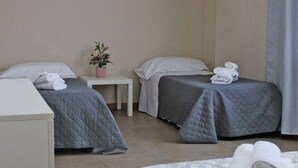 A hotel room with two beds, each with a grey quilted bedspread and white pillows. There is a small white table with a pink flower pot between the beds. - 03. Apartment (Signa)