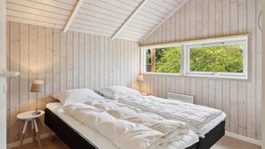 A bedroom with a sloped ceiling, wooden walls, a large window with a view of greenery, a bed with white bedding, and a bedside table with a lamp. - Amazing home in Sjællands Odde (Sjællands Odde)