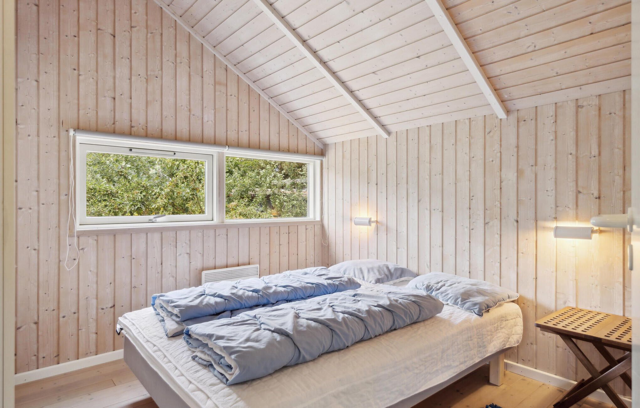 A bedroom with a sloped ceiling, wooden walls, a bed with blue bedding, a window with a view of greenery, and a small wooden stool.