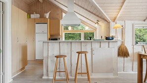 A modern kitchen with wooden cabinets, a central island with stools, a microwave, and a view of greenery outside. - Amazing home in Sjællands Odde (Sjællands Odde)