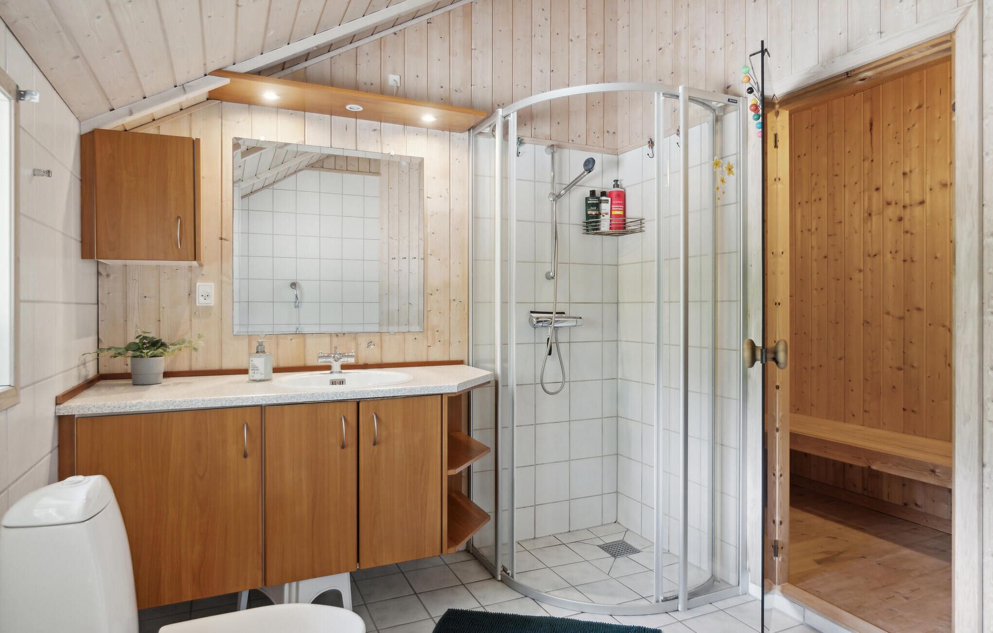 A modern bathroom with a wooden vanity, a large mirror, a shower enclosure, and a small plant.