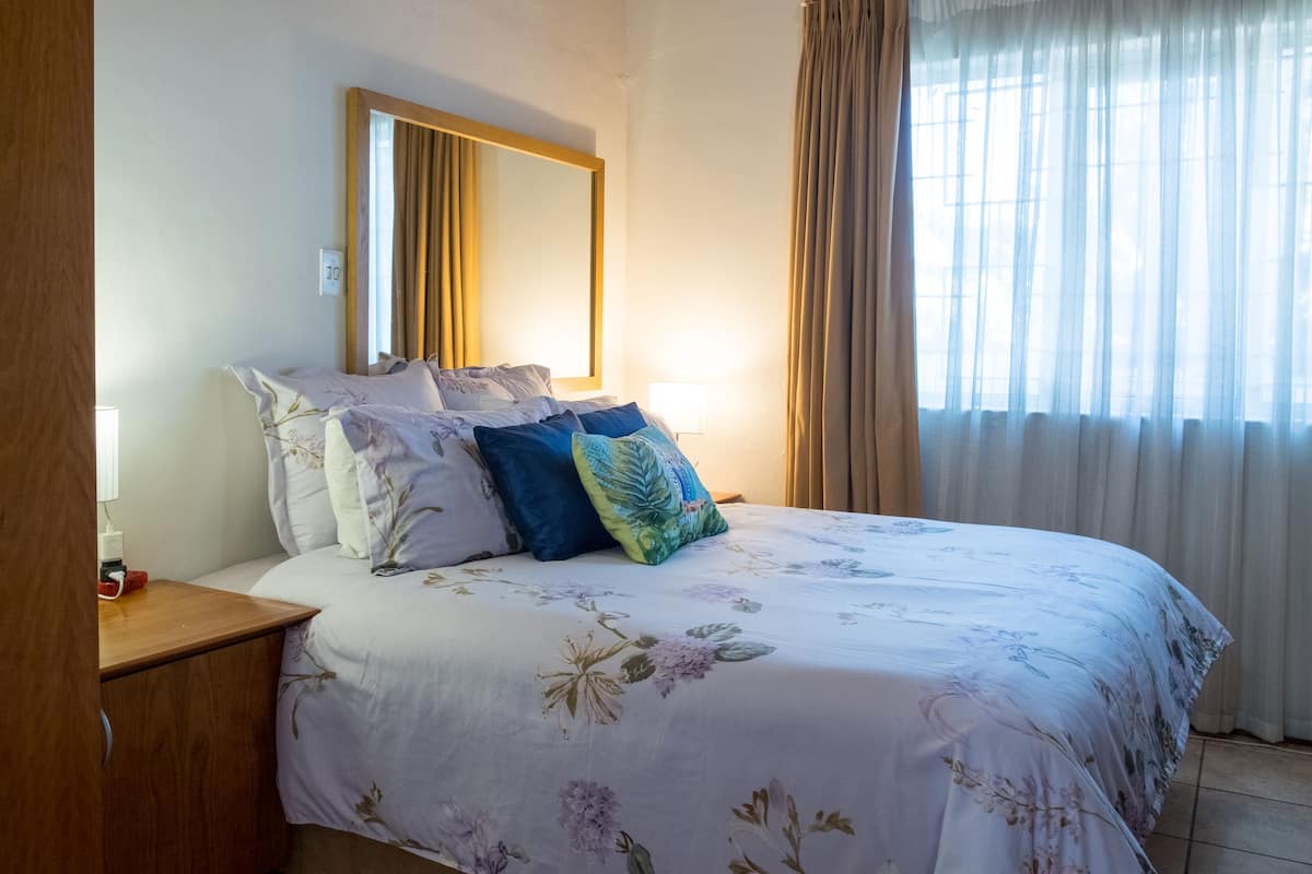 A bedroom with a bed, a bedside table, a mirror, and a window with curtains.