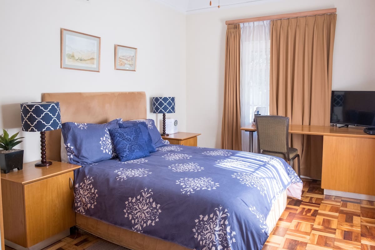 A bedroom with a bed, two bedside tables, a desk, a chair, a television, and two framed pictures on the wall.