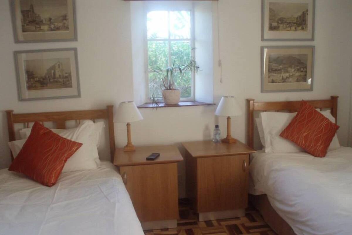 A hotel room with two beds, a small wooden nightstand with lamps, a window with a plant, and framed pictures on the wall.