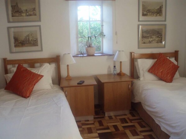 A hotel room with two beds, a small wooden nightstand with lamps, a window with a plant, and framed pictures on the wall.