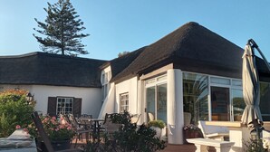 Ein Haus mit einem Strohdach, weißen Außenwänden und großen Fenstern. Draußen stehen Möbel und Blumentöpfe.