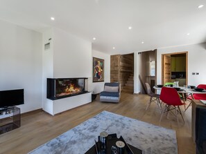 Uma sala de estar moderna com lareira, uma televisão de tela plana, uma área de jantar com cadeiras vermelhas e uma cozinha visível por uma porta aberta.