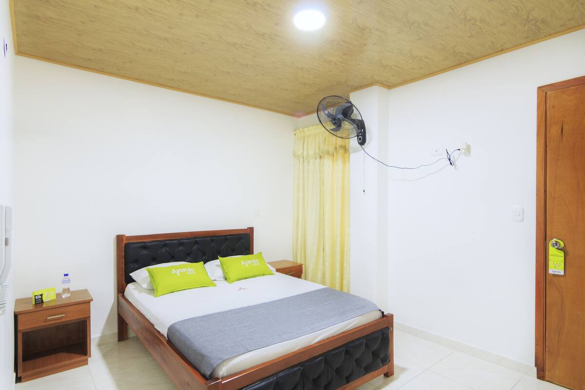 Une chambre d’hôtel avec un lit en bois, une table de chevet, une commode, un ventilateur fixé au mur et une porte avec une serrure.