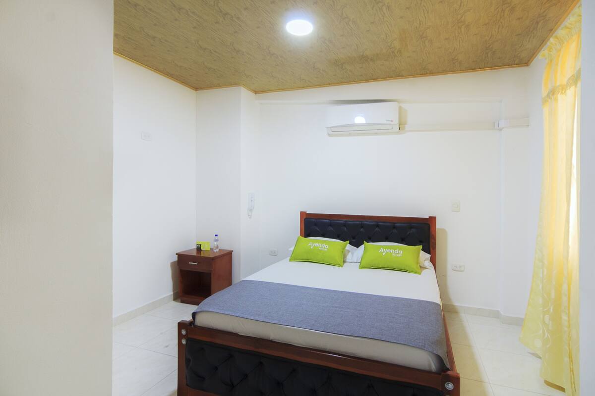 Une chambre à coucher comprenant un lit, une table de chevet, un appareil de climatisation et une fenêtre avec des rideaux.