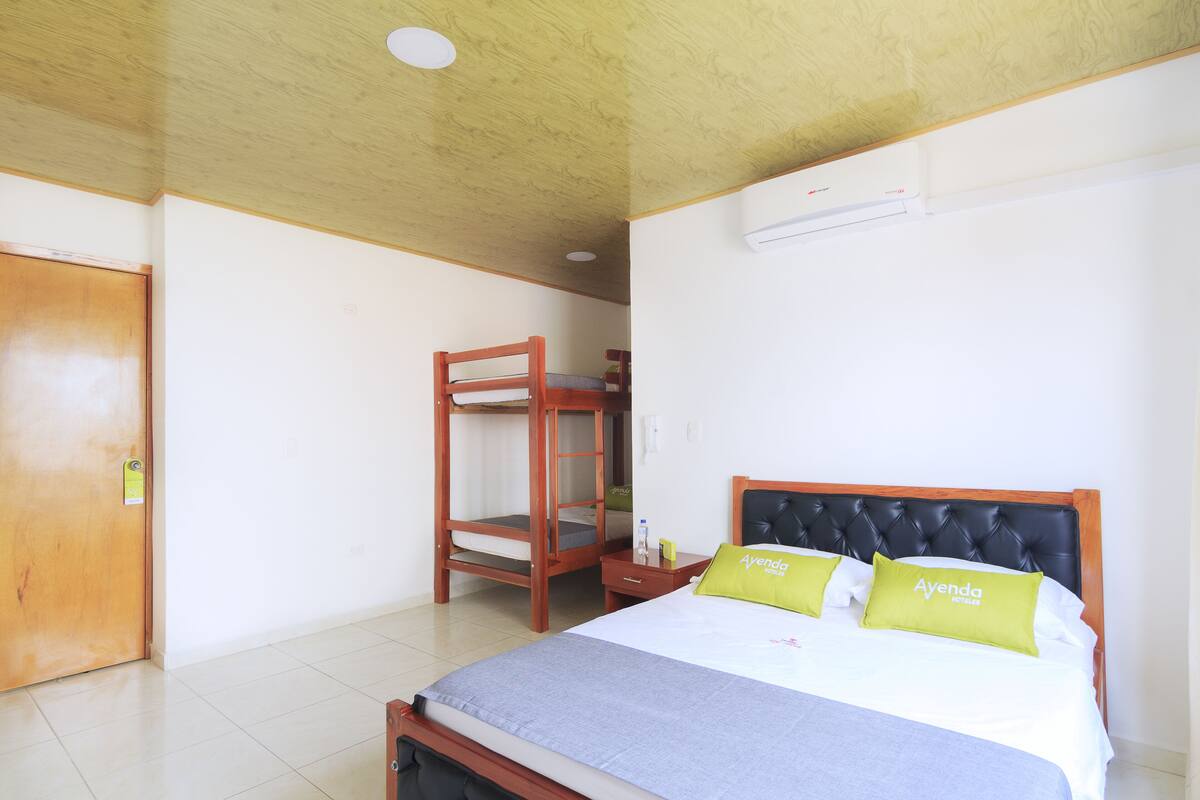 Une chambre d’hôtel avec un lit superposé, un lit simple avec une tête de lit, une porte en bois et un appareil de climatisation blanc fixé au mur.