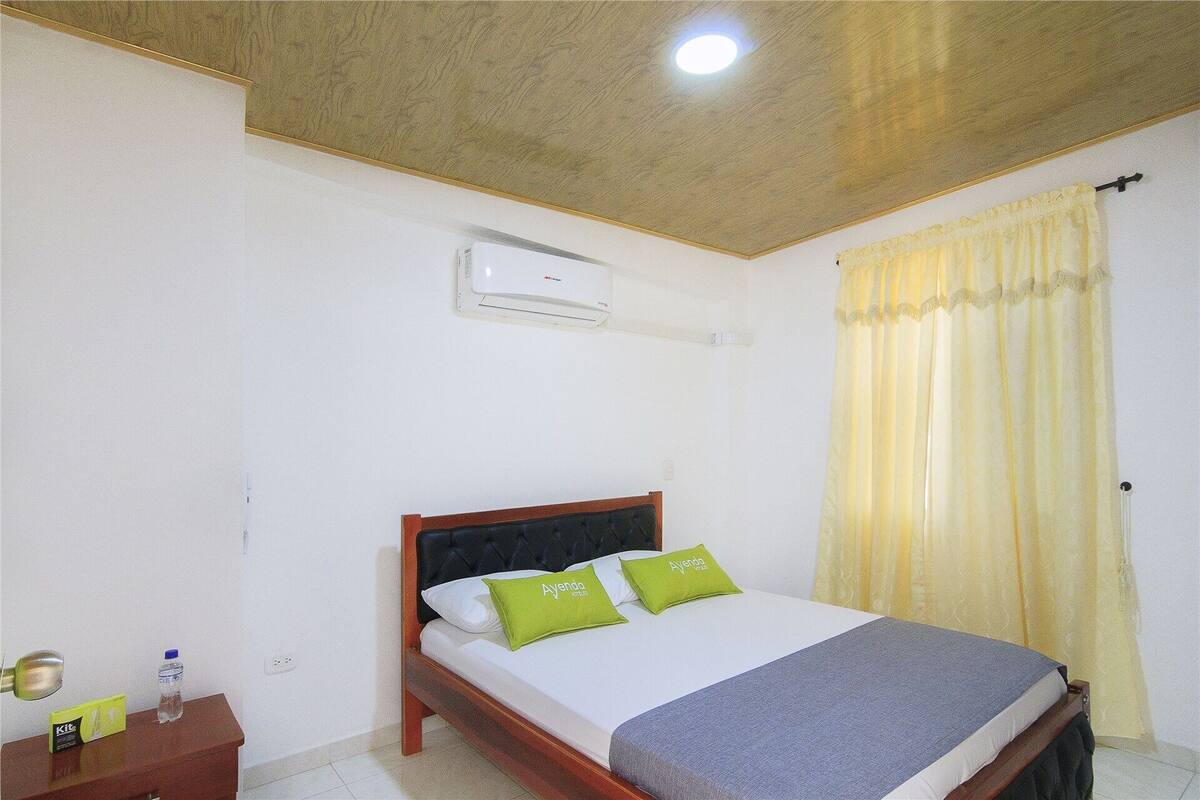 Une chambre d’hôtel comprenant un lit, une table de chevet, un appareil de climatisation et une fenêtre avec des rideaux.