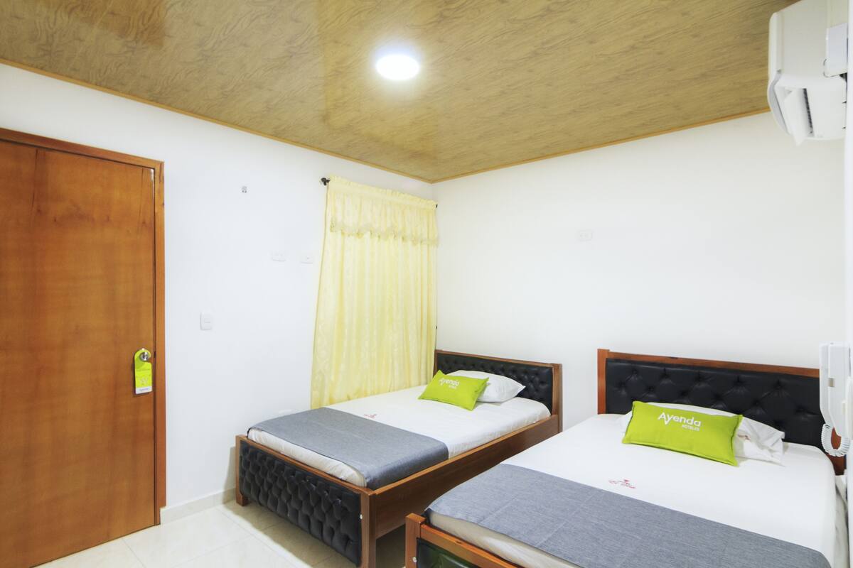 Une chambre avec deux lits, des têtes de lit en bois, une porte en bois, un rideau jaune et un plafonnier blanc.