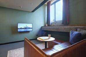 Una habitación con un sofá, una mesa con un libro y un vaso, un televisor en la pared y una ventana con cortinas.