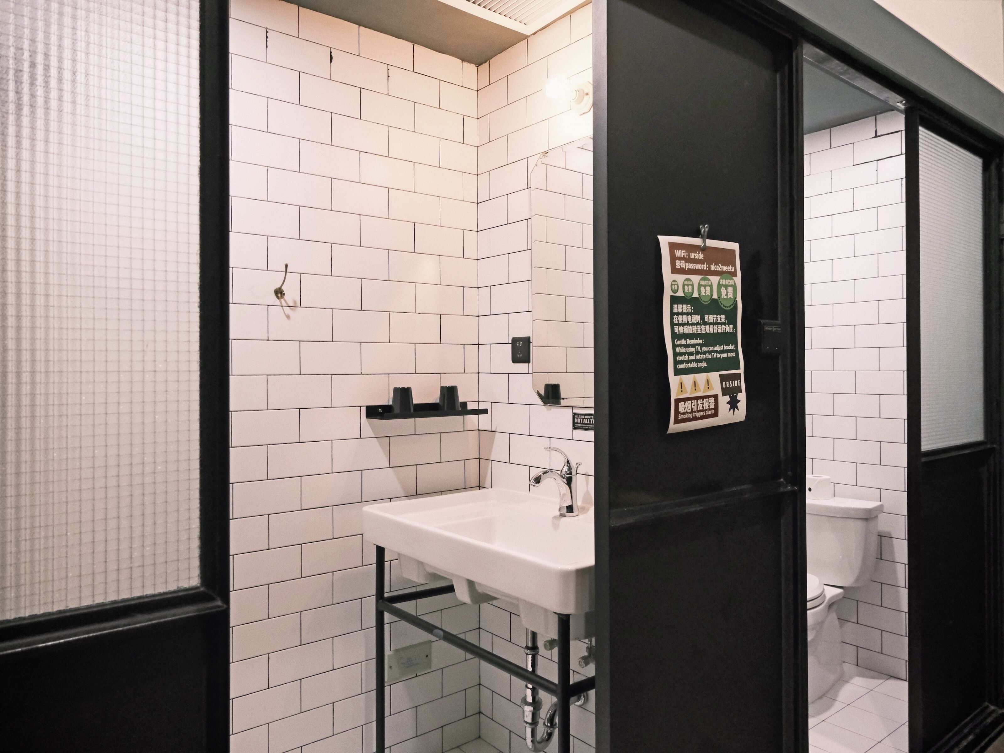 A bathroom with white tiled walls, a black door, and a white sink with a black shelf above it.