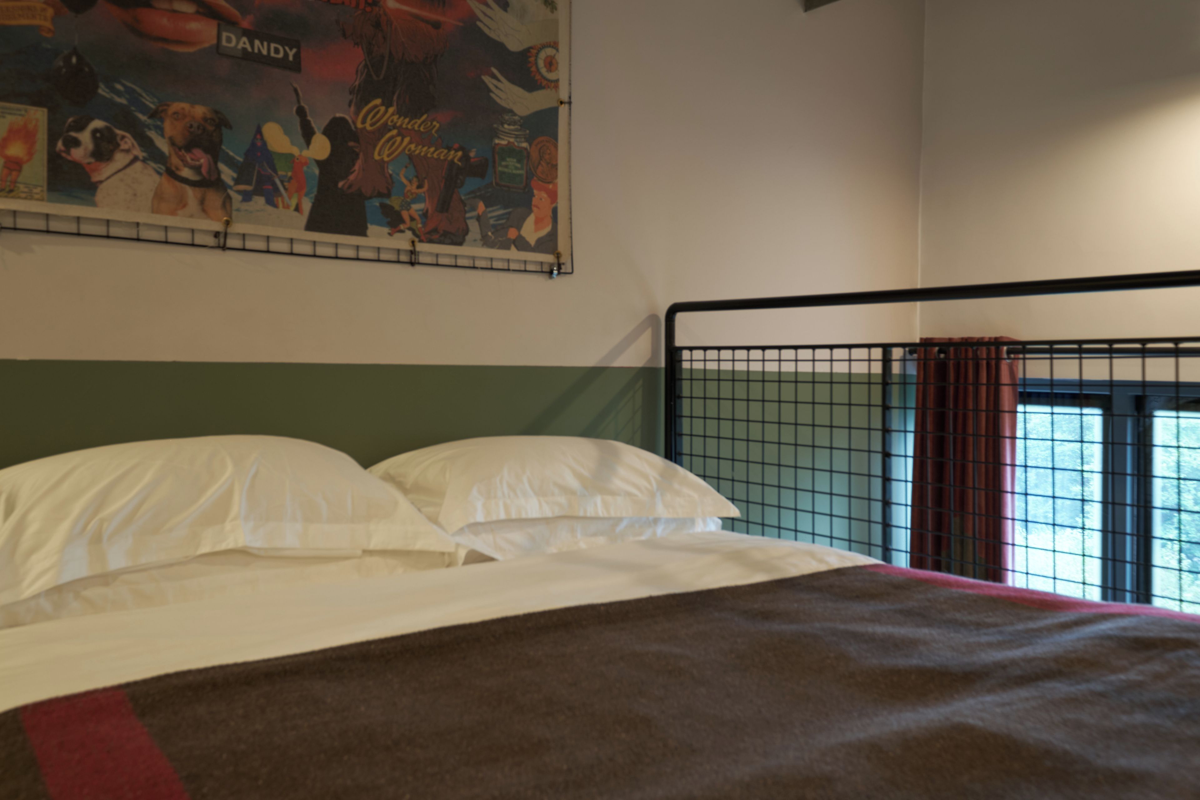 A bed with white linens and a brown blanket, a wall with a colorful poster, and a window with a metal grid.