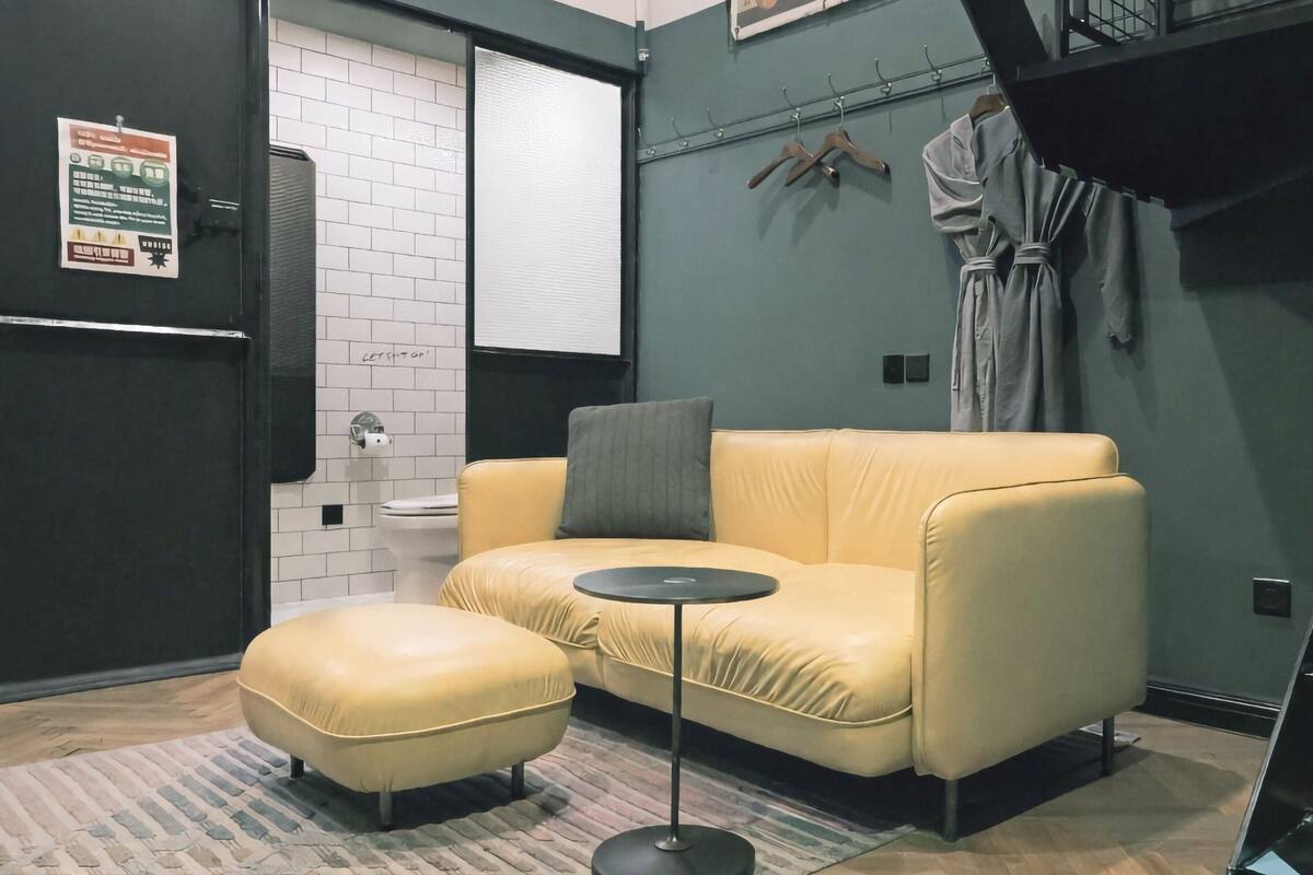 Een moderne kamer met een gele bank, een klein rond tafeltje, een kamerkleed met patroon, een spiegel met zwarte lijst en een wit toilet.