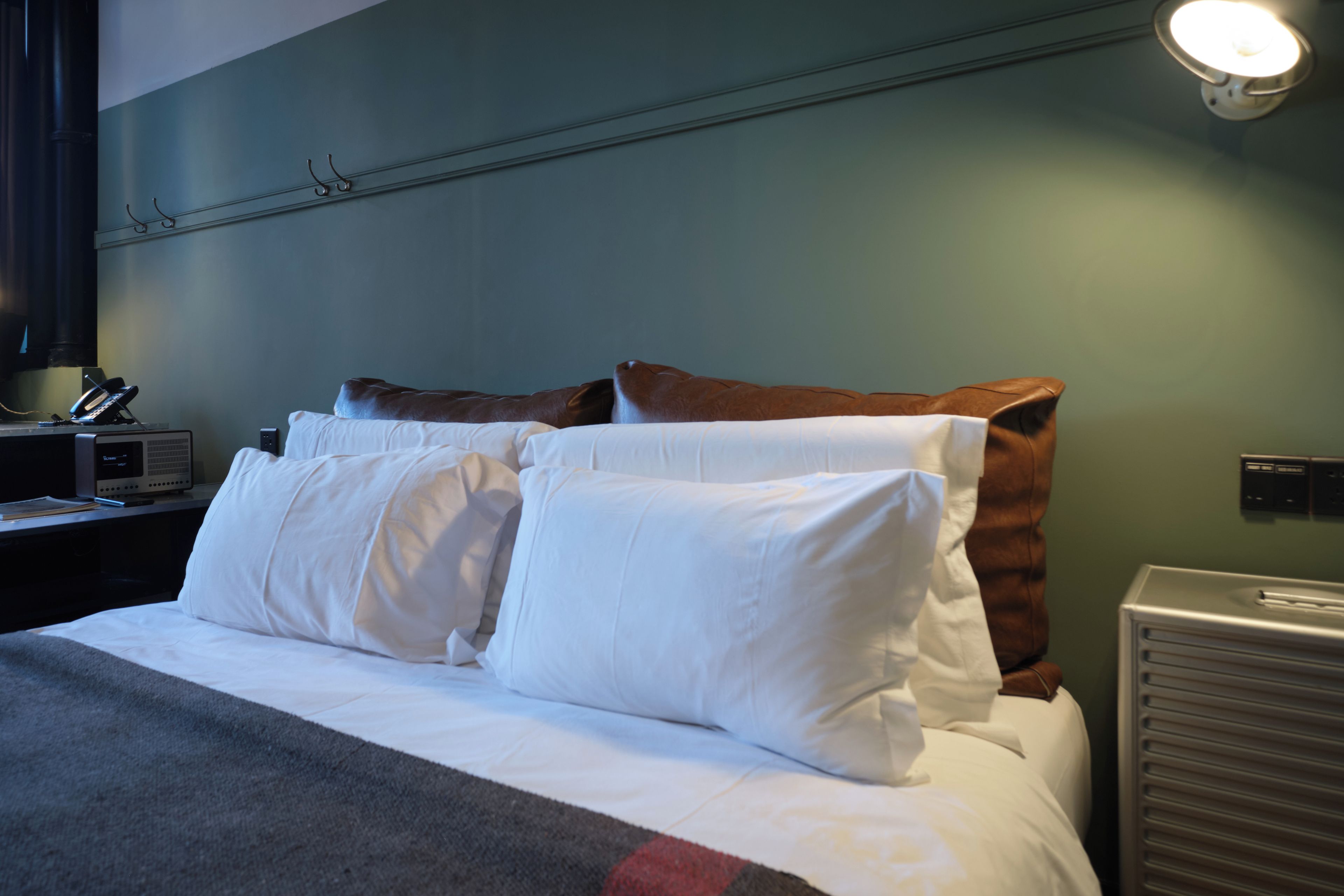 Una cama bien hecha con sábanas blancas y un cabecero marrón, una mesita de noche con una lámpara, y una lámpara de pared.